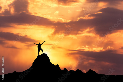 Landscape with silhouette of a standing happy man with backpack and raised-up arms on the mountain peak on the background of cloudy sky at colorful sunset in summer. Travel, Climbing, Trekking.