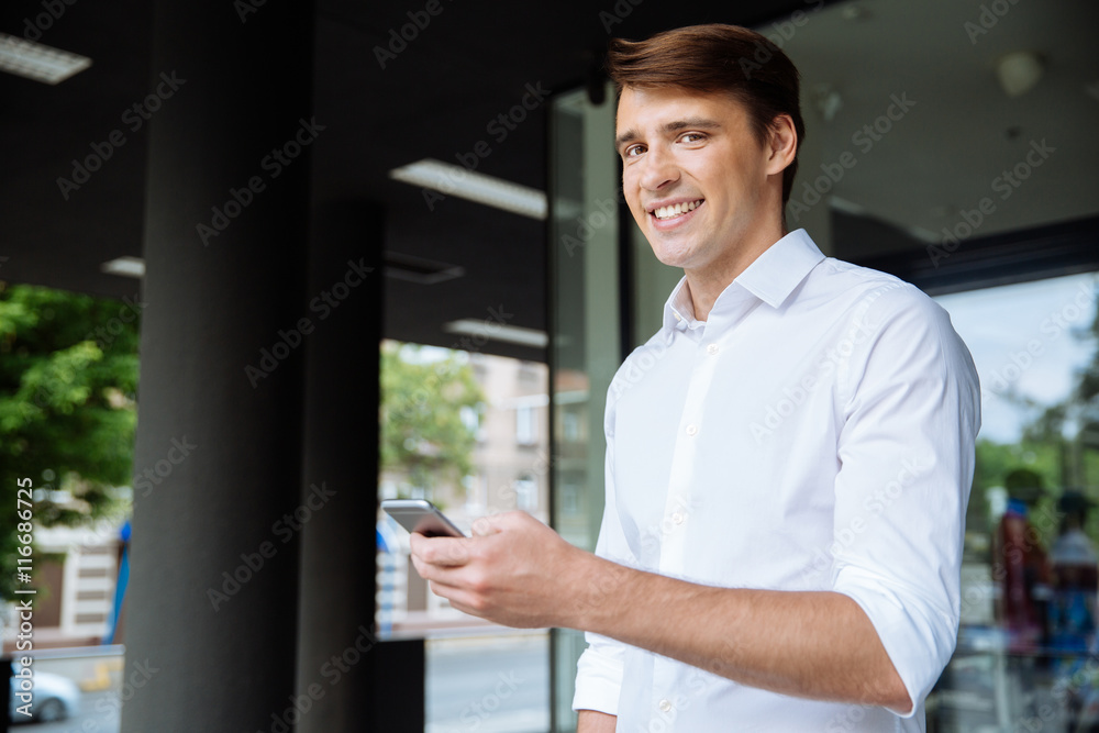 Cheerful young businessman using smarphone and smiling near business center Cheerful young businessman using smarphone and smiling near business center