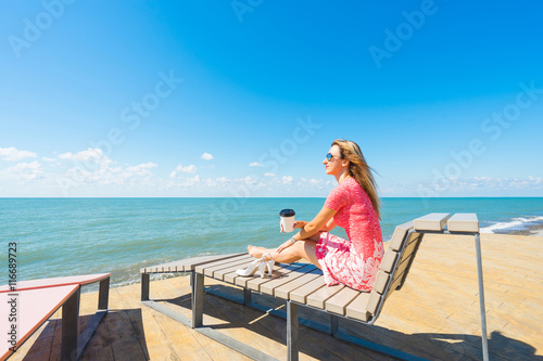 Young beautiful woman in red dress  sitting on beach chair with shoes, drinking coffee and looking on blue sea and sky