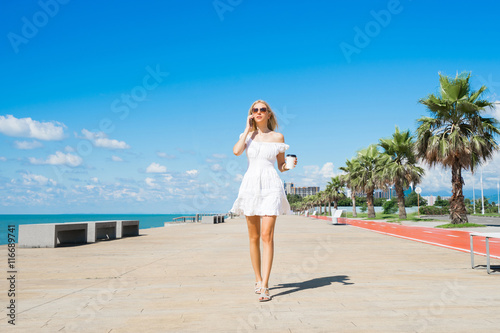 Young  beautiful woman in white dress walking on the beach boulevard with coffee cup and talking on mobile.