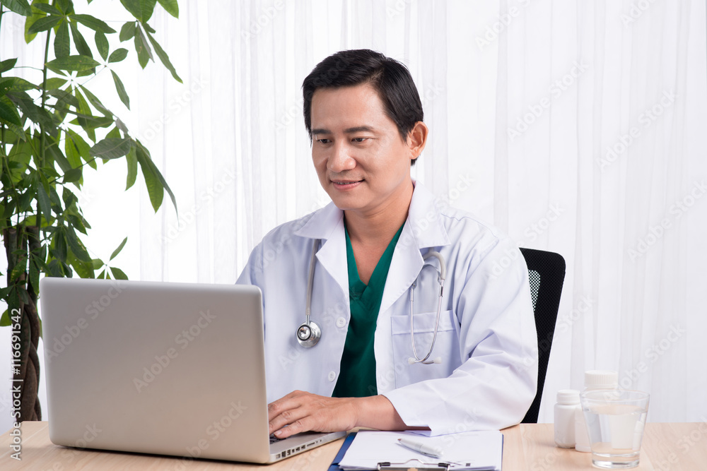 Smiling doctor working on computer at his desk in medical office Smiling doctor working on computer at his desk in medical office