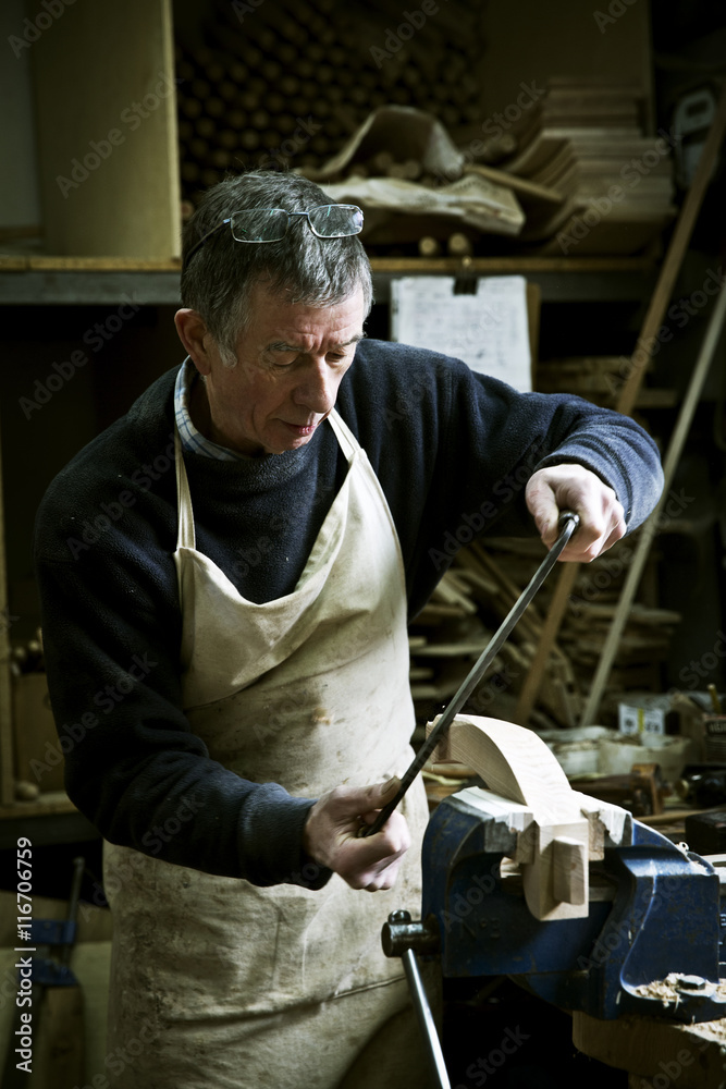 A man working in a furniture maker's workshop using a rasp on a wooden shape in a clamp. A man working in a furniture maker's workshop using a rasp on a wooden shape in a clamp.