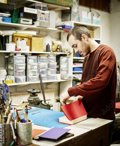 A man working in a book binding workshop, creating a red cover for freshly stitched pages. 