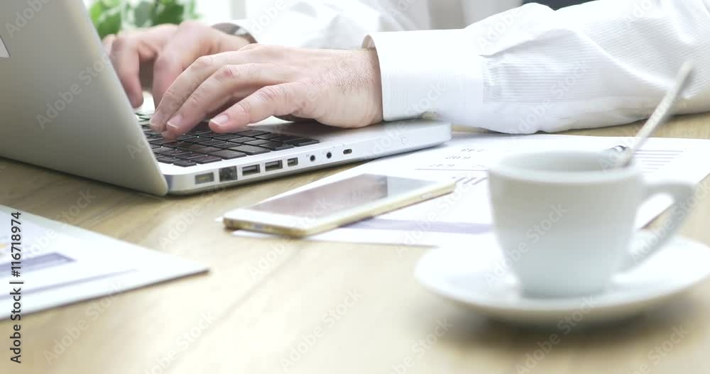 Corporate businessman working at office desk, he is typing with a laptop, cup of coffee on foreground Corporate businessman working at office desk, he is typing with a laptop, cup of coffee on foreground