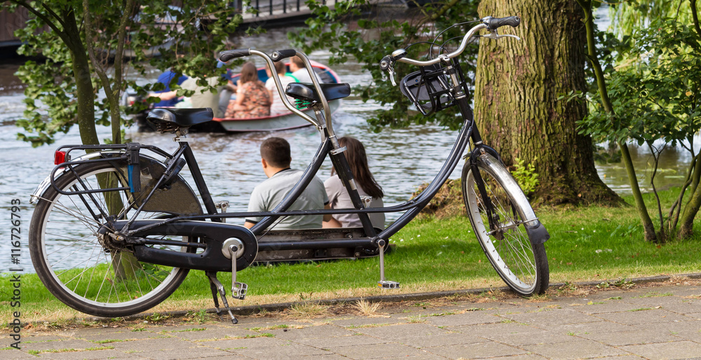 The tandem bicycle and young couple relaxing near the water.