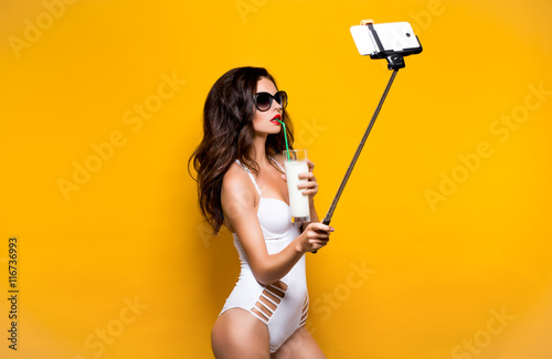 Beautiful brunette model in sunglasses and white monokini taking selfie with cell on selfiestick while drinking cocktail. Studio shot photo
