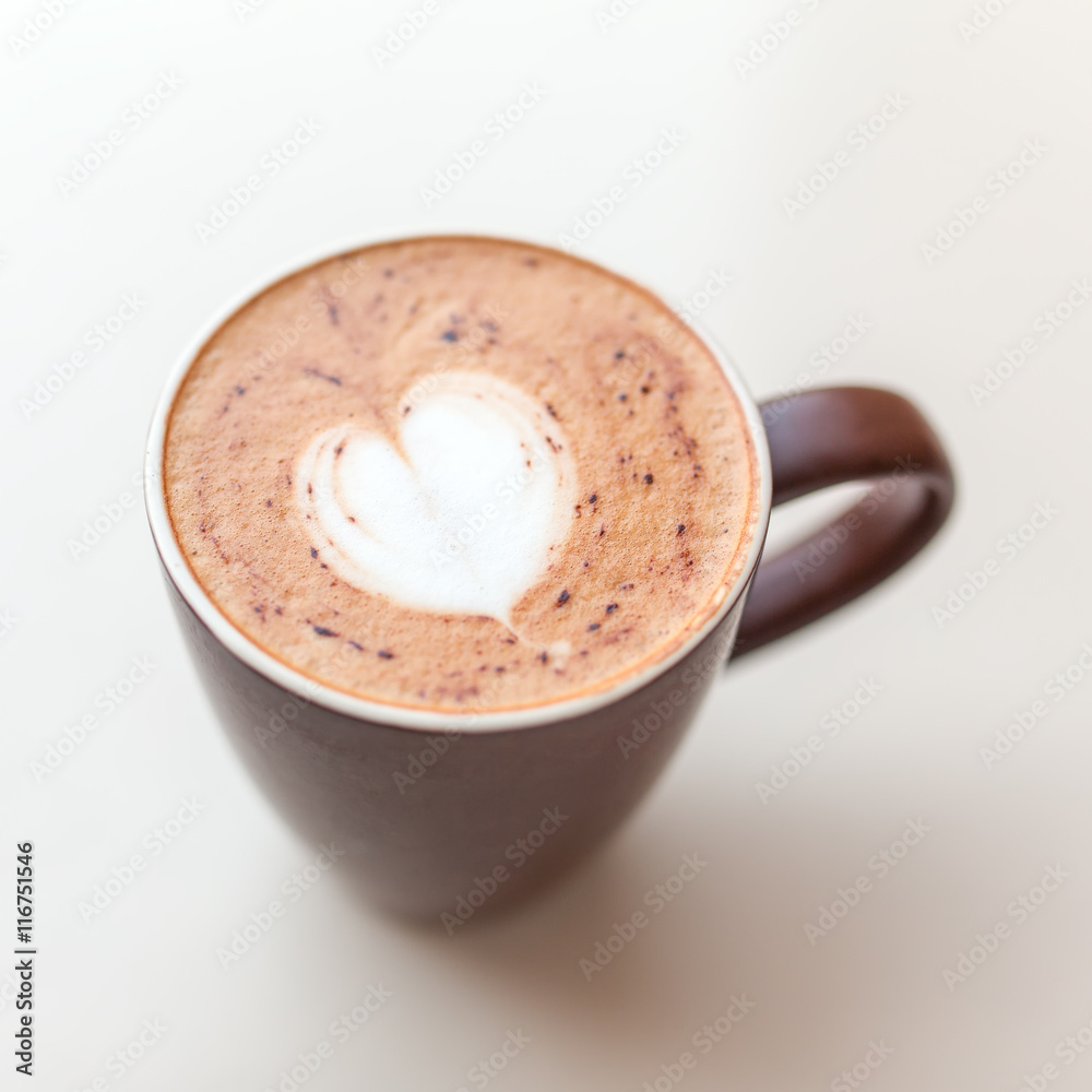 Cup of cappuccino coffee with the milky foam on top in a heart shape viewed from above in a brown cup on white table Cup of cappuccino coffee with the milky foam on top in a heart shape viewed from above in a brown cup on white table