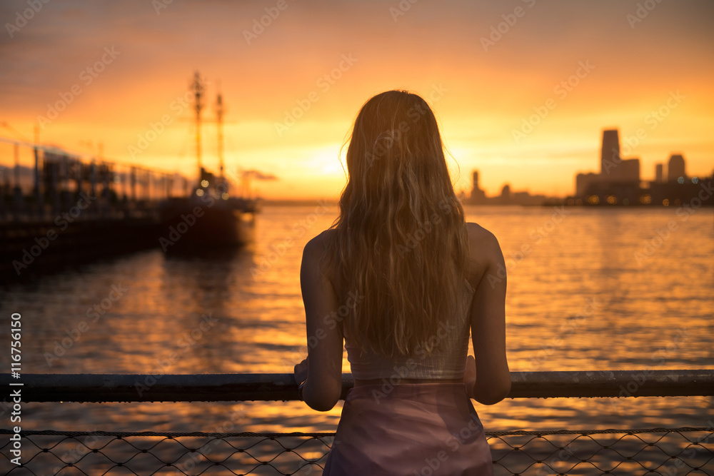 Beautiful girl standing on ocean pier with yacht and looking at sunset sky. Girl from the back outdoors near the ocean.
