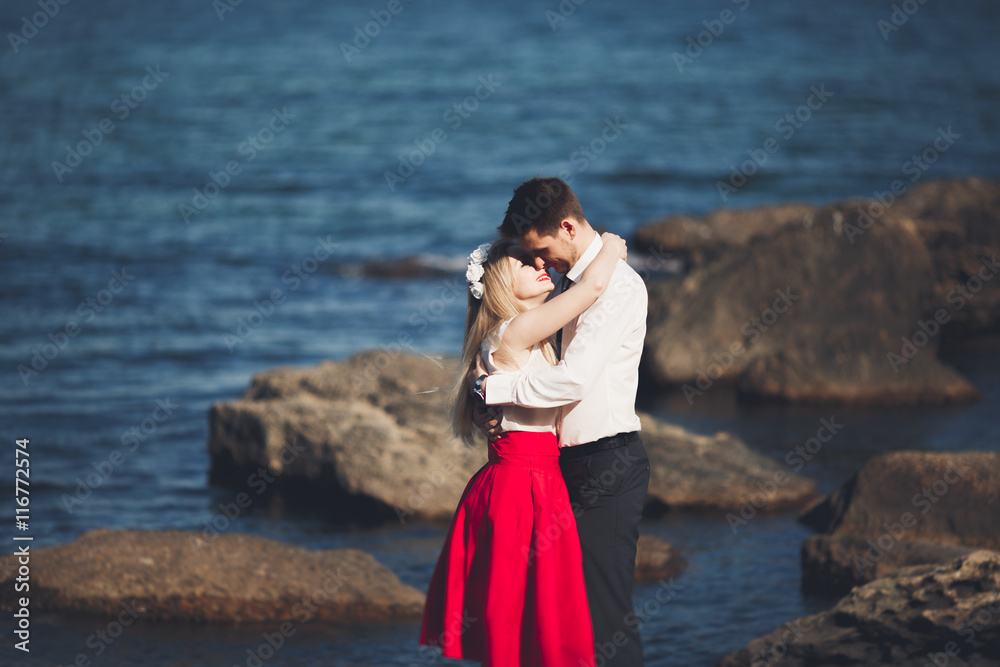 Romantic loving couple posing on stones near sea, blue sky Romantic loving couple posing on stones near sea, blue sky
