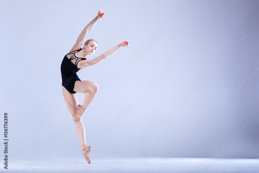 Naklejka premium Ballet Perfection. Young and beautiful ballerina in a black dancing suit is posing in a white studio full of light.