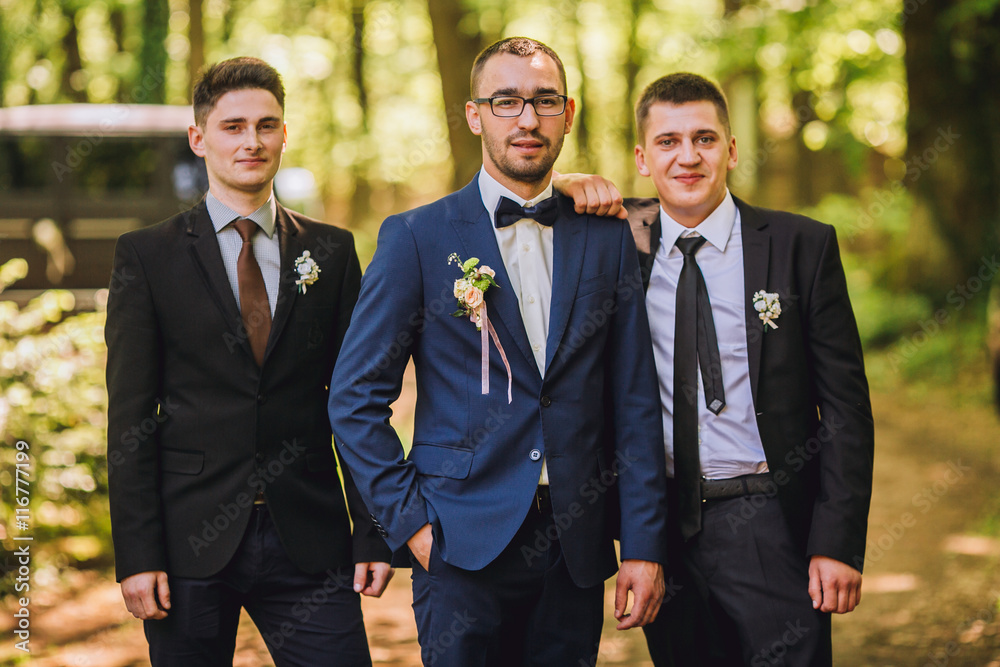 Confident smiling handsome groom in black suit with two groomsman in pine wooden park Confident smiling handsome groom in black suit with two groomsman in pine wooden park
