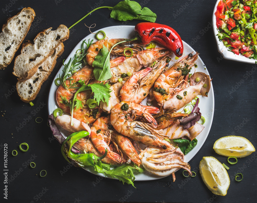 Plate of roasted tiger prawns and octopus with fresh leek, vegetable salad, peppers, lemon, bread and pesto sauce over black background, top view, horizontal composition Plate of roasted tiger prawns and octopus with fresh leek, vegetable salad, peppers, lemon, bread and pesto sauce over black background, top view, horizontal composition