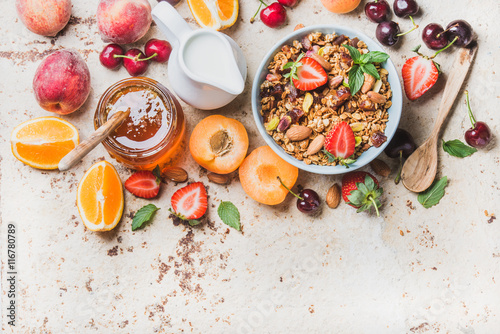Healthy breakfast ingredients. Oat granola in bowl with nuts  strawberry and mint leaves  milk in pitcher  honey in glass jar  fresh fruits  berries and mint on light concrete background  top view