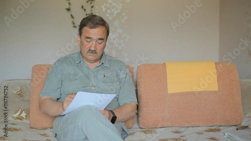 Charming male senior citizen signing a legal document. A man reads the document carefully and sign each page. Houses on the sofa