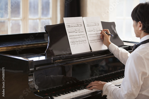 Young man sitting at a grand piano in a rehearsal studio, annotating sheet music.