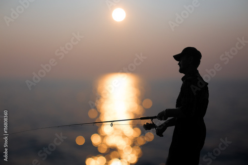 fisherman at dusk. silhouette of a fisherman with a fishing rod in the background reflected in water sunset.
