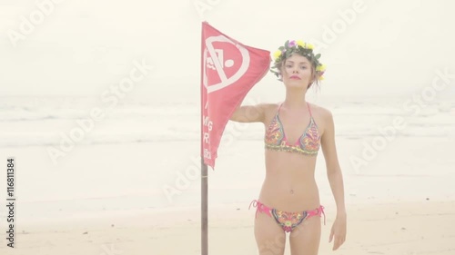 Beautiful blonde glamour woman wearing colourful swimwear and wreath smiling and posing with red warninig flag on a beautiful summer day at the beach over sea and sky background photo