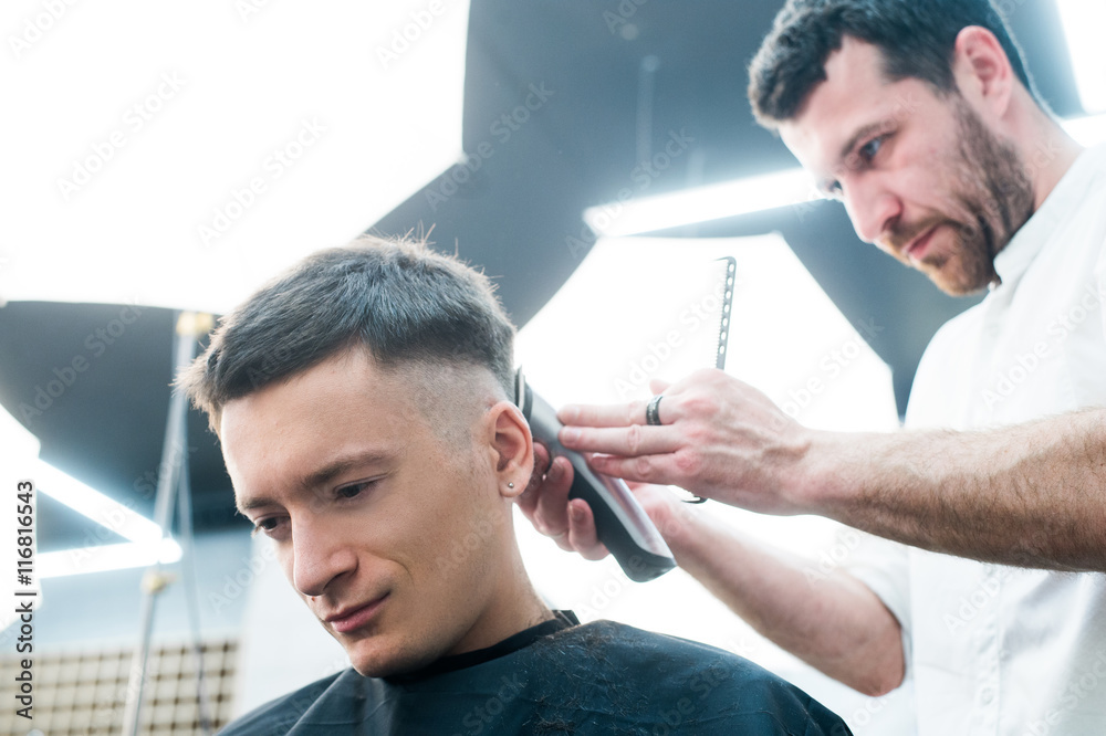 Fototapeta premium Professional styling. Close up side view of young man getting haircut by hairdresser with electric razor at barbershop