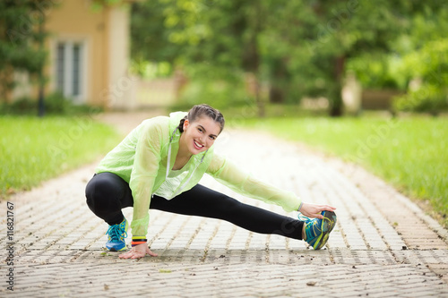 Fototapeta Naklejka Na Ścianę i Meble -  Smiling young woman is exercising outdoors. She is doing stretching exercise for her legs.