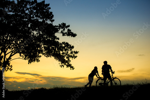 sillhouette of man and woman walking park with vintage bicycle at sunset time.