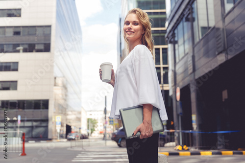 Beautiful young businesswoman with coffee holding laptop on city background © snedorez