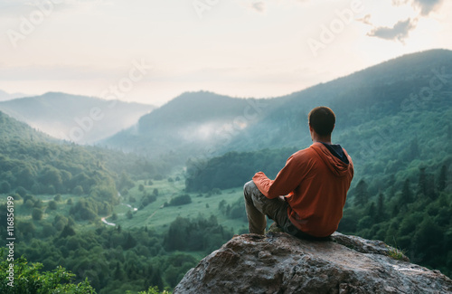 Traveler young man sitting on stone in the summer mountains and enjoying view of nature