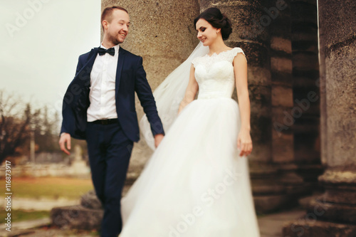 Groom holds bride's hand walking with her from the ruined cathed photo
