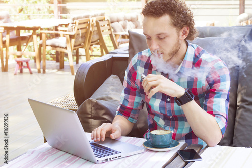 Stylish vapor sitting in cafe. Modern handsome man with laptop outdoor