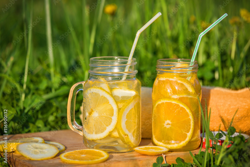 Two glass jars with lemonade on a wooden stand on the grass. on the Board the slices of lemon and orange. Terry towel orange Two glass jars with lemonade on a wooden stand on the grass. on the Board the slices of lemon and orange. Terry towel orange