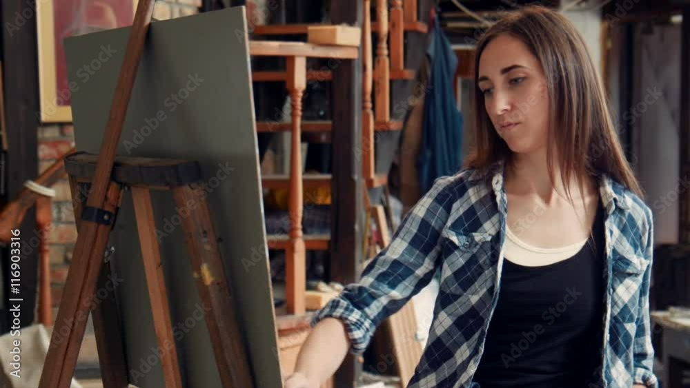 Young woman artist standing painting a canvas in a studio with her head tilted to the side and an absorbed expression Young woman artist standing painting a canvas in a studio with her head tilted to the side and an absorbed expression
