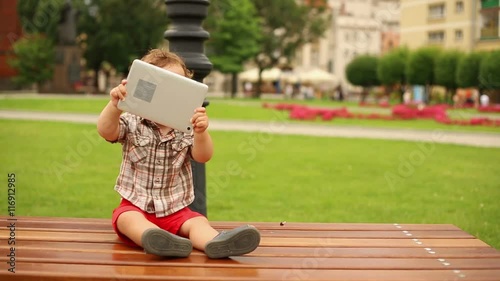 Cute kid is sitting on a bench with tablet 
