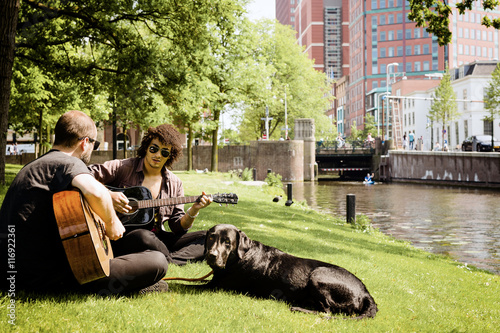 Two young men playing acoustic guitar in a city park in the company of a black dog. Selective focus.