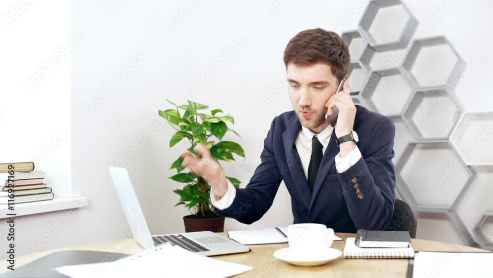 Young successful businessman speaking on phone at his workplace in office. Slow motion. Young successful businessman speaking on phone at his workplace in office. Slow motion.