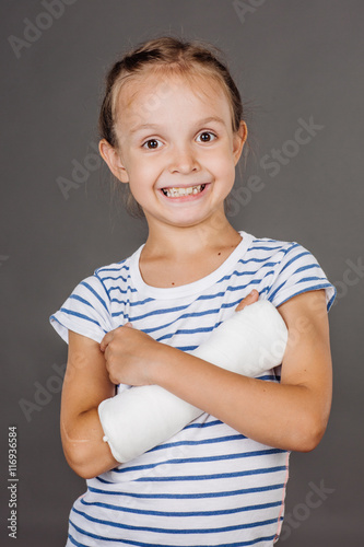 Happy smiling girl with broken arm is standing on the gray background.