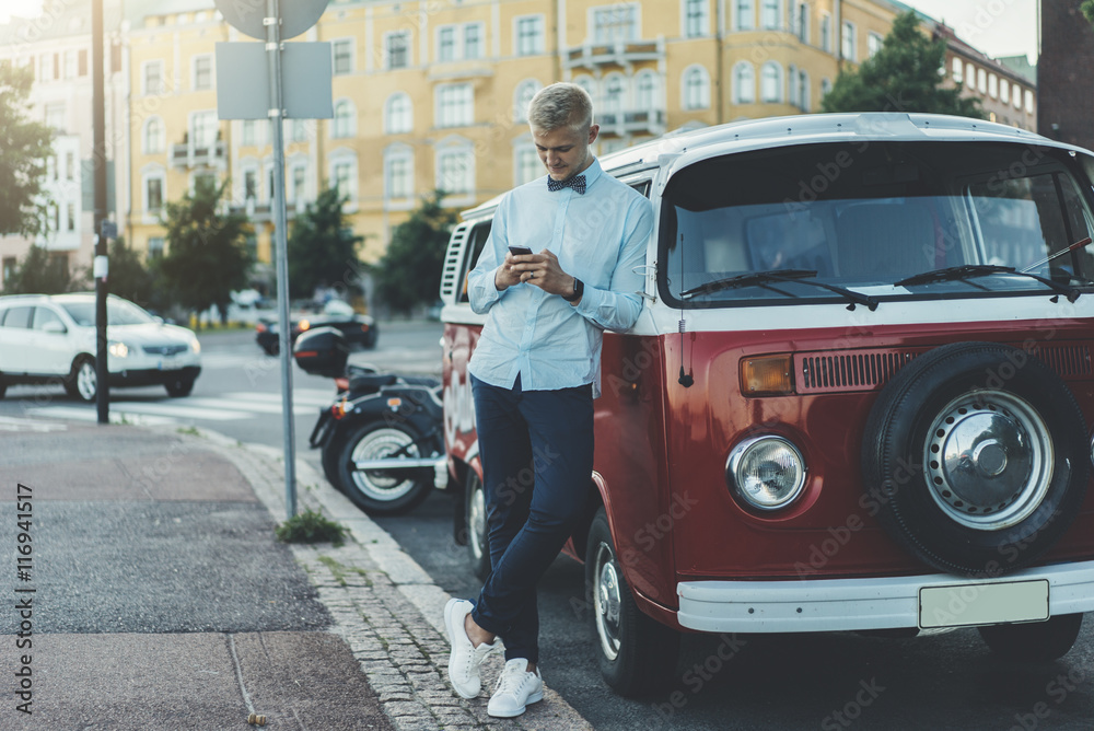 Fototapeta premium Young happy smiling man wearing blue shirt with bowtie and using modern smartphone outside, hipster guy typing text message or reading information via cellphone