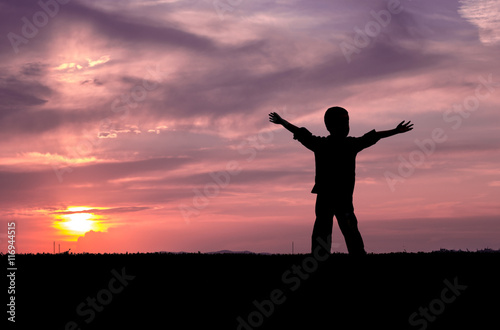 Child silhouette on sunset with raised hands looking into the sun and dramatic sky with clouds. Concept of happiness, freedom, youth, timeless, serenity, comfort, joy, childhood memories.
