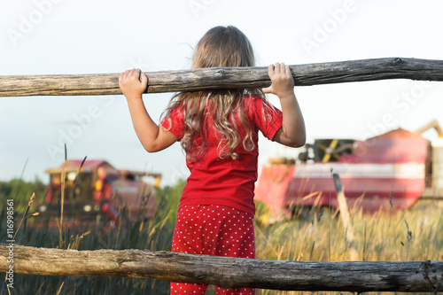 Curious farm girl wearing polka dot kids pans looking at field with working red combine harvesters