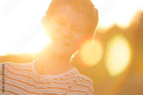 Headshot portrait of screwing up eyes laughing teenage boy in warm sunset backlit with lens flares