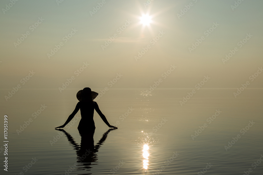 Silhouette. Young woman with hat on head enjoy the sunset on the beach and she feels the harmony in herself. Serene and warm, gentle water. Touch to water. In front of view opens infinity.