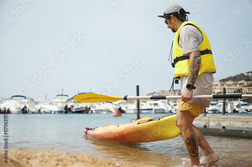 Half profile full-length portrait of young strong Caucasian man on vacation, pulling kayak out from the water, standing against sand beach, with tattooed hands and legs. Leisure activities and hobbies