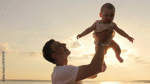 Happy man playing lifting his daughter outdoors. Silhouette of young father holding up his cute baby daughter in air against sunset sky playfully