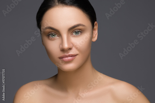 Pretty girl with big eyes and dark eyebrows,  naked shoulders, looking at camera  smiling, a model  light nude make-up, gray studio background, beauty photo, copy space, close . © Alexander Molofeev