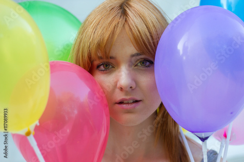 Portrait of a white girl with orange hair sitting on a white stairs holding colorful balloons with their hands. 