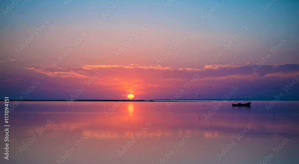 A remote boat in the quiet sea  on picturesque sunset background. Beautiful reflection of sun and sky in the calm water. Evening glow. Outdoor shot. Copy-space