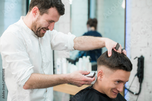 Professional styling. Close up side view of young satisfied man getting haircut by hairdresser with electric razor at barbershop