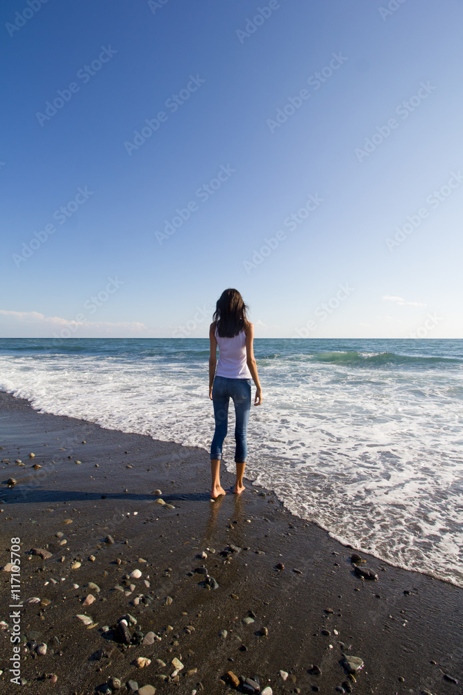 Beautiful young happy girl wearing white T-shirt and blue jeans playing with the waves on a beach.