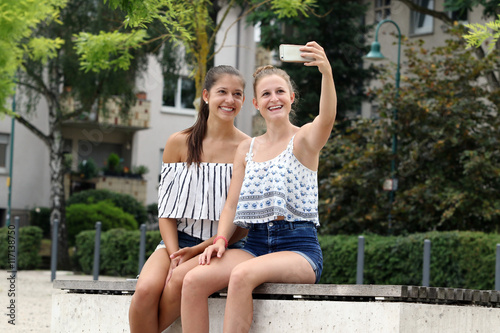 Lachende junge Frauen sitzen auf einer Bank im Park und machen ein Selfie mit ihrem Smartphone