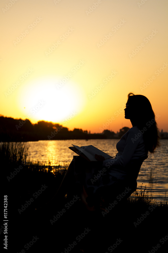 outlines of a beautiful young woman with a book by the river at dawn of the day