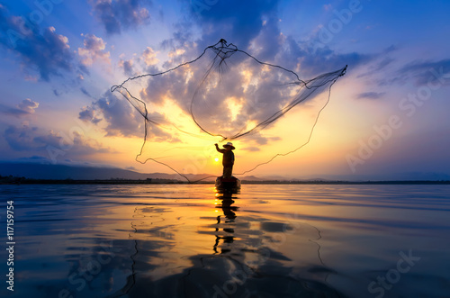 Fotografie, Tablou Asian fisherman on wooden boat casting a net for catching freshwater fish in nat