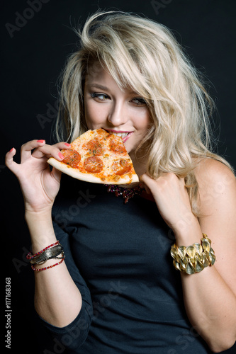 Young happy beautiful woman eating pizza. Pretty young sexy woman in black dress with long blonde hair and makeup holding tasty big slice of pizza ready to eat. Standing in studio on black background